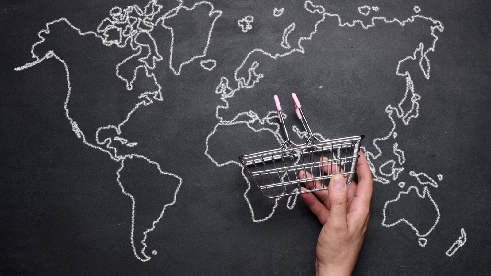 A female hand holding a miniature empty shopping basket against the background of a drawn world map. Concept of online shopping, international shipping